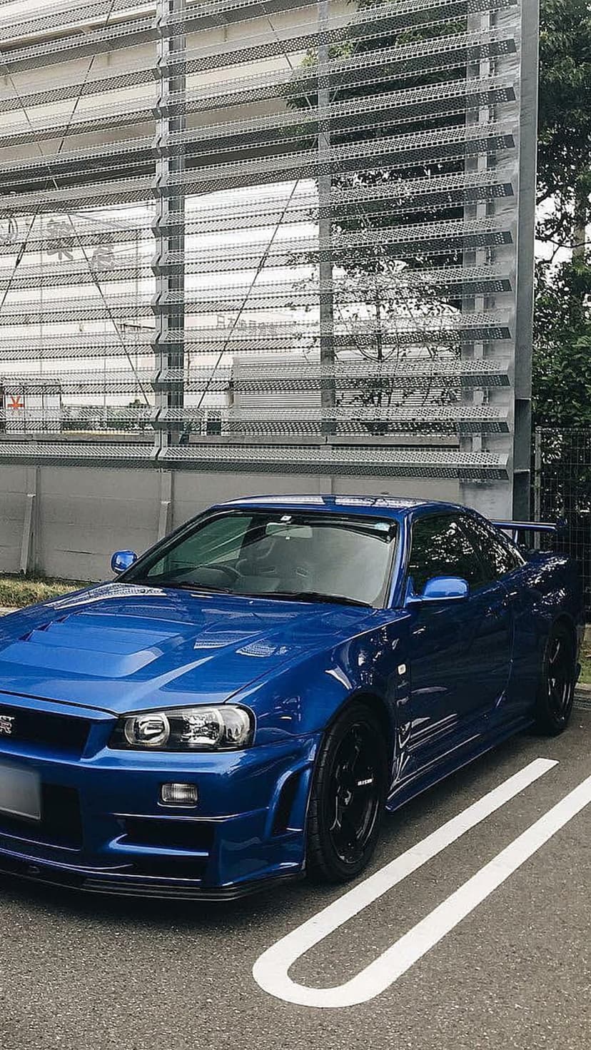Blue Nissan Skyline R34 GT-R with black wheels parked in front of a modern slatted metal building.