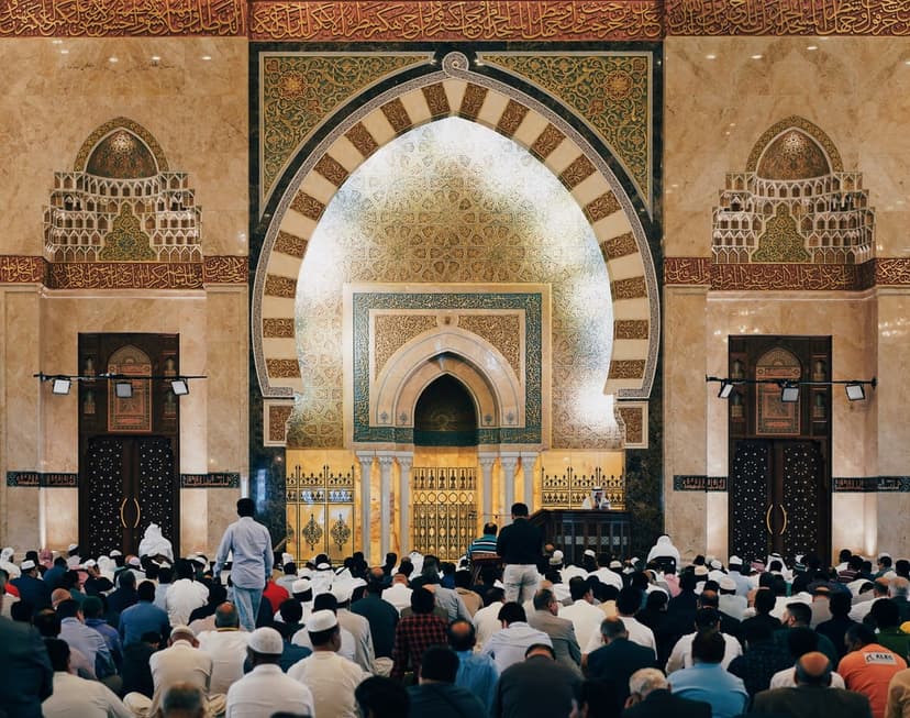 Men seated on a carpeted floor inside a grand mosque featuring ornate arched doorways and intricate tilework.