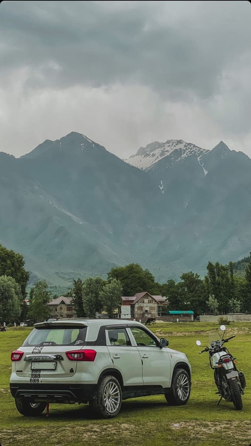 White SUV and black motorcycle parked in a green meadow with snowy mountains and pine trees in the background.