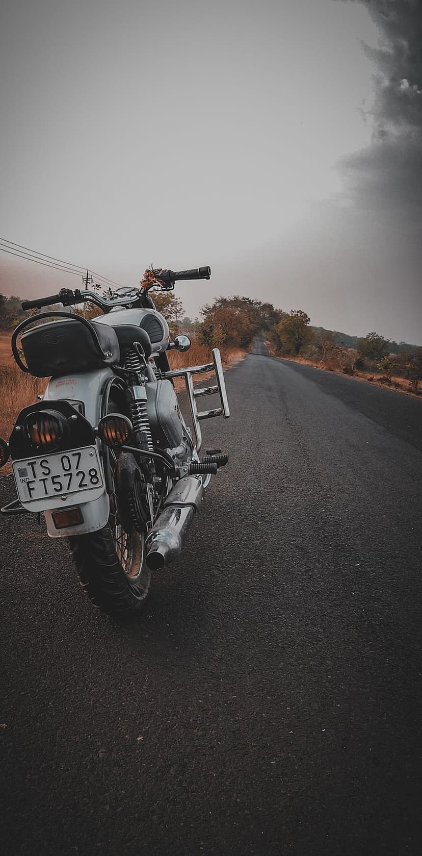 Royal Enfield motorcycle parked on an empty rural road under a dark overcast sky with dry autumn vegetation.