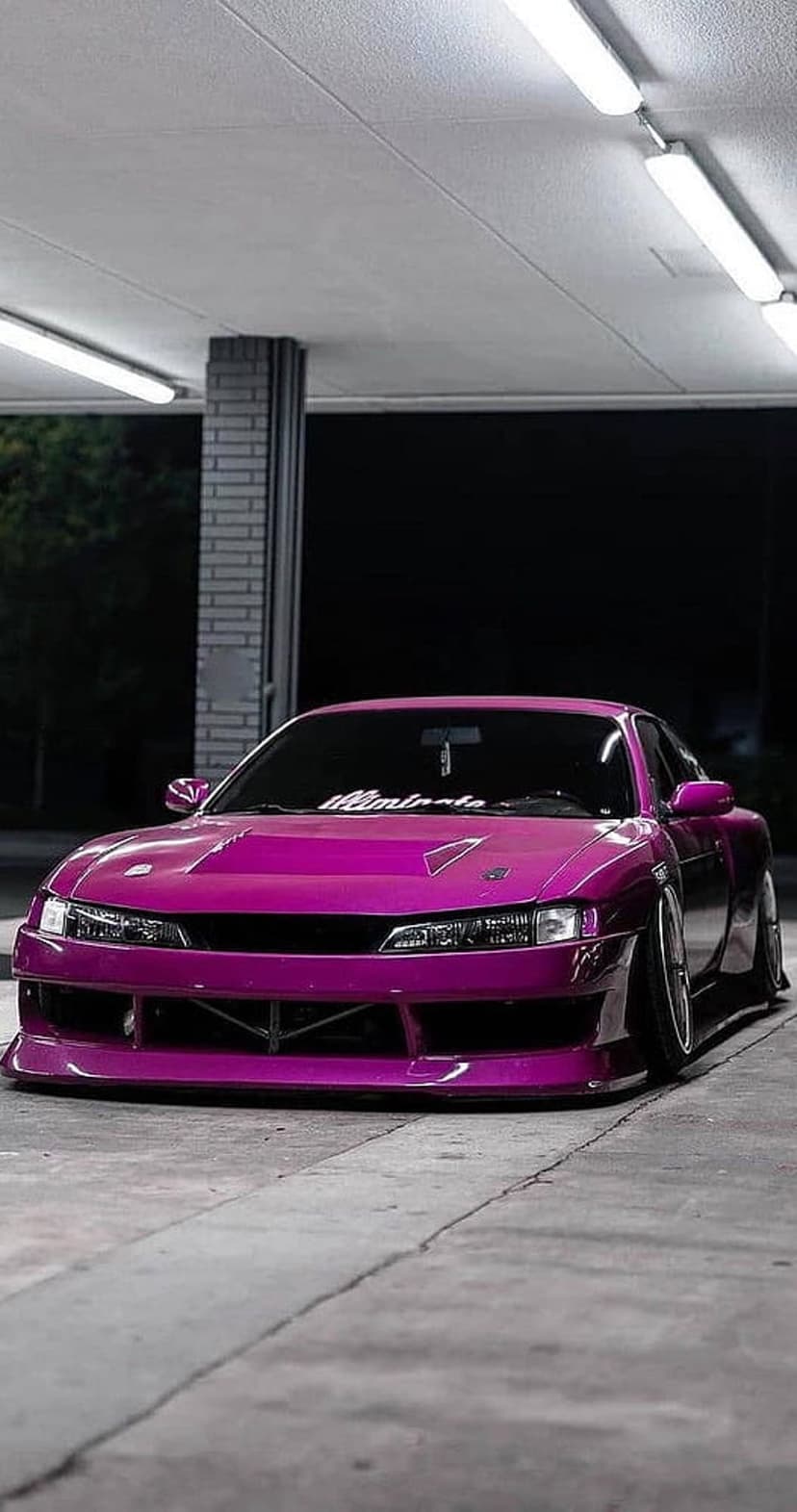 Pink Nissan Silvia S14 with a widebody kit and slammed suspension parked under bright fluorescent garage lights.