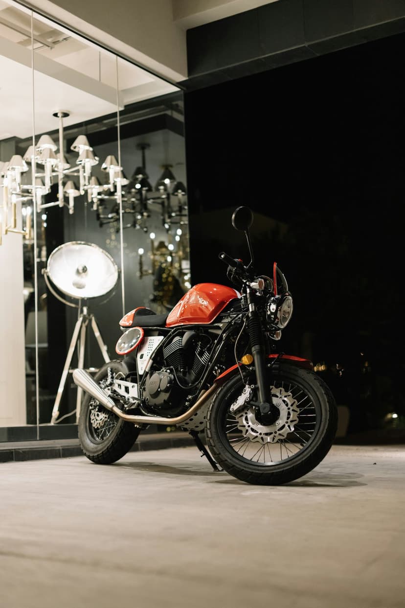 Red cafe racer motorcycle parked on a light floor with a vintage spotlight and chandelier reflection behind it.