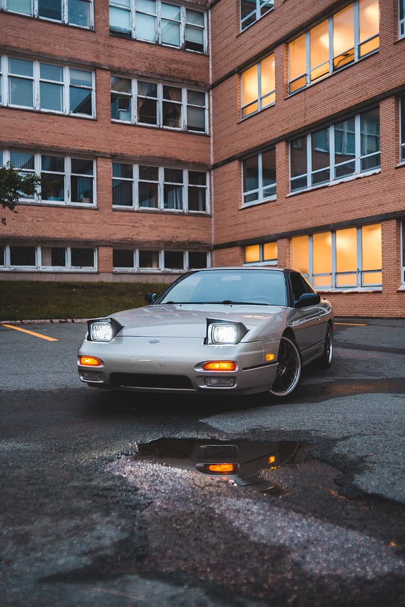 Nissan 240SX with pop-up headlights on wet asphalt before a brick building with glowing windows at night.