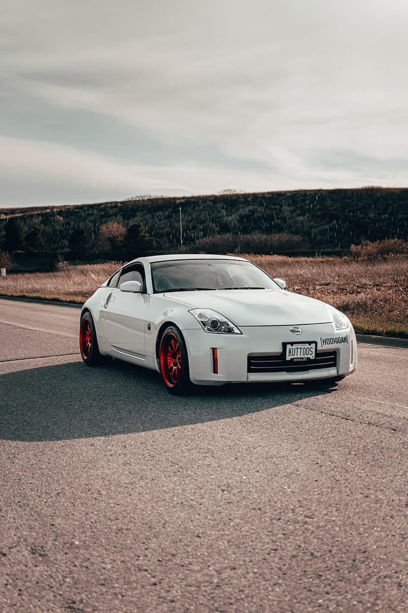 White Nissan 350Z with red wheels parked on a paved road with hills and trees under a cloudy sky in the distance.