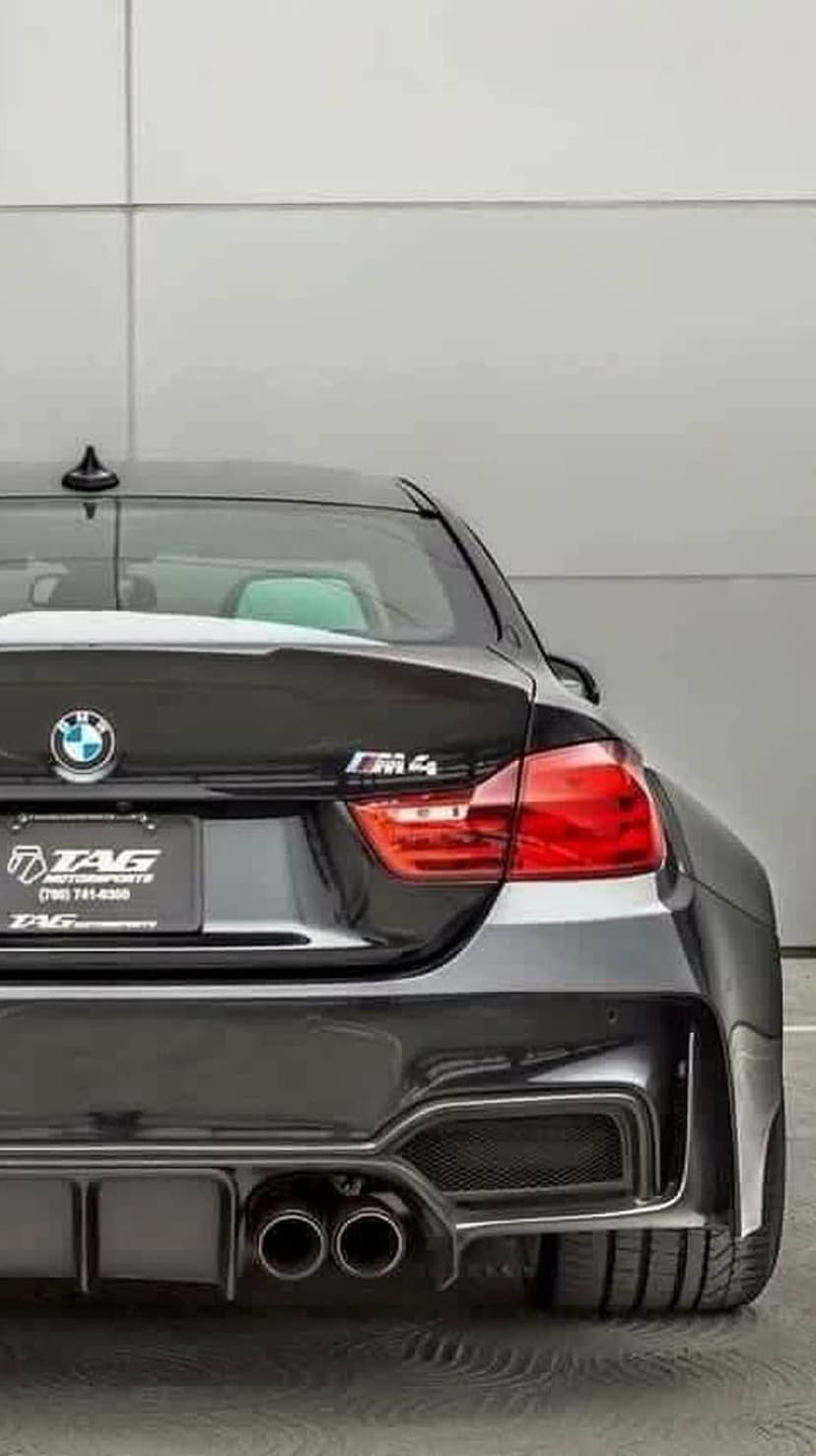 Black BMW M4 rear view showing red taillights, quad exhaust, and M4 badge against a white concrete garage wall.