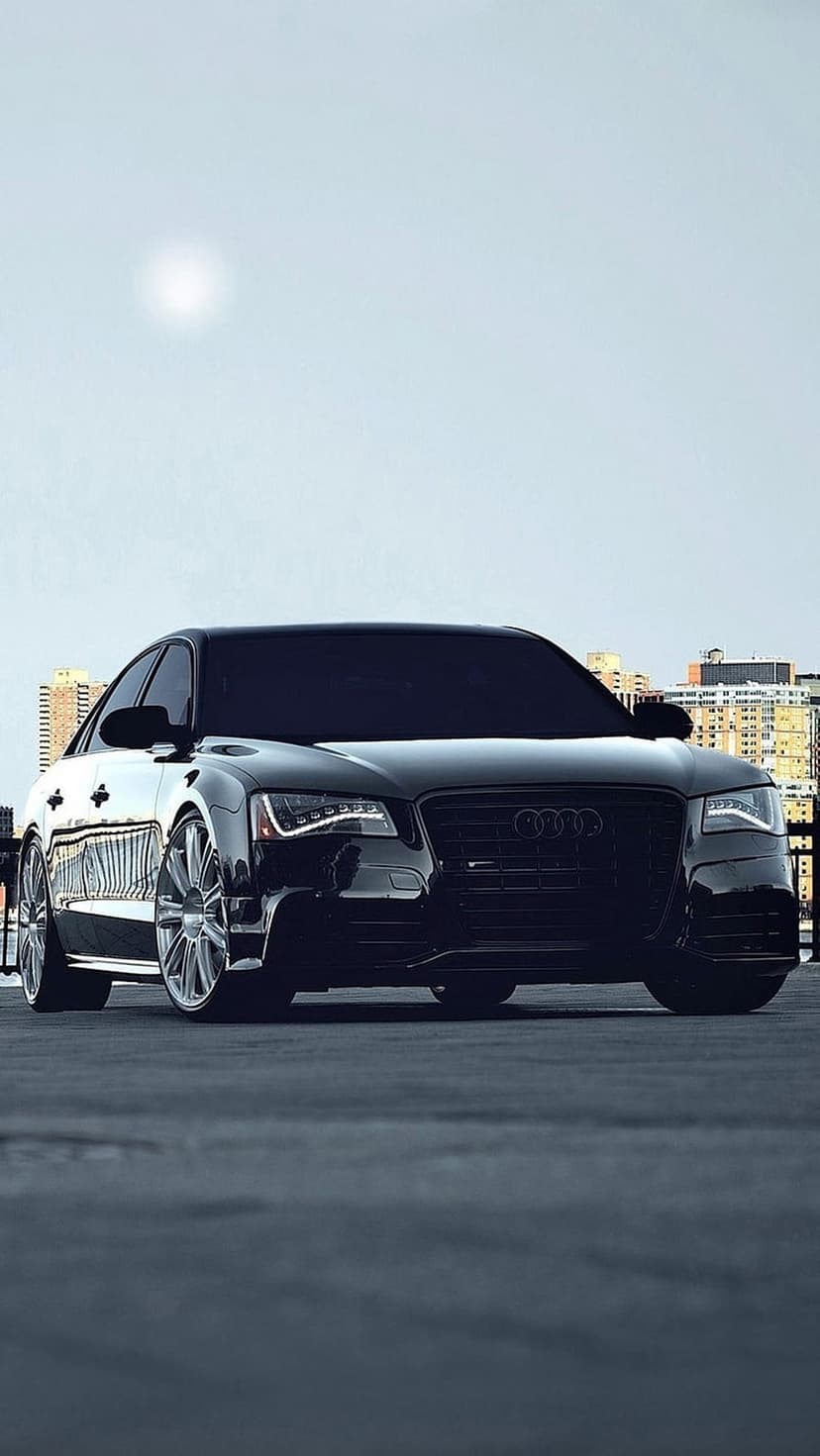 Black Audi luxury sedan parked on asphalt with a blurred metropolitan skyline and bright sky in the background.