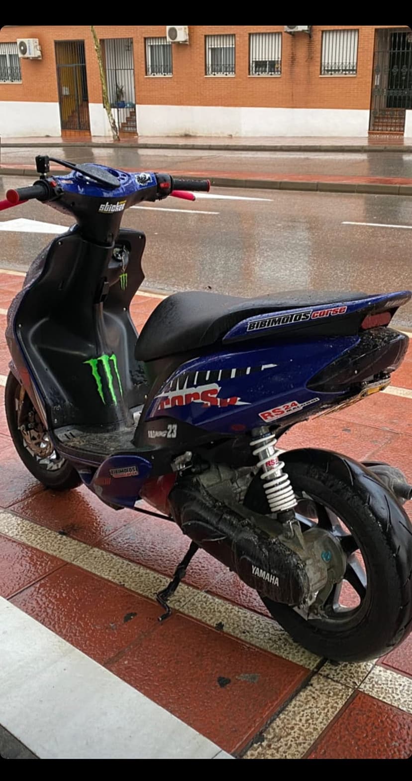 Yamaha scooter with Monster Energy graphics and red grips parked on a wet brick sidewalk after rain.