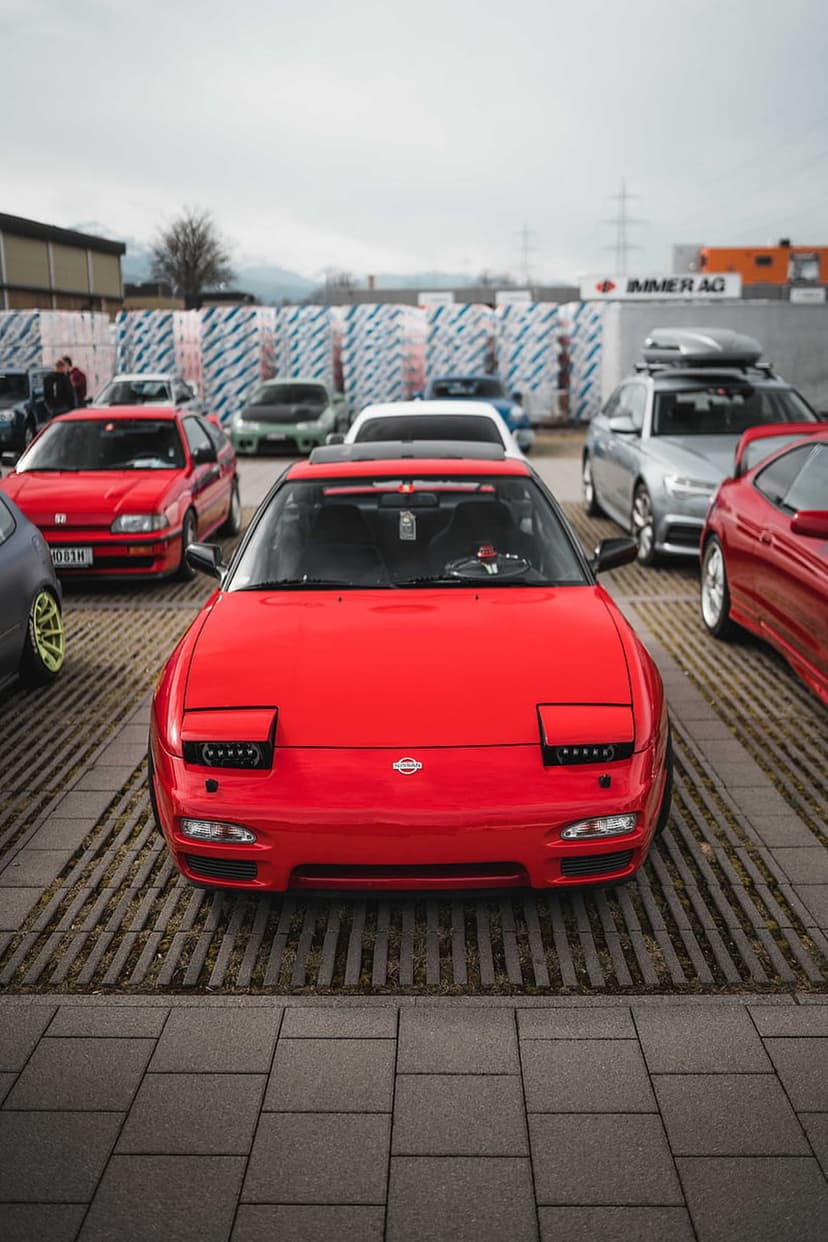 Red Nissan 240SX S13 with pop-up headlights parked at an outdoor car show on a sunny day with blurred background.
