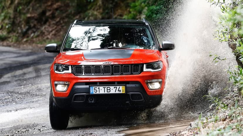 Red Jeep Compass Trailhawk driving through a deep puddle with a large water spray on a scenic wooded trail.