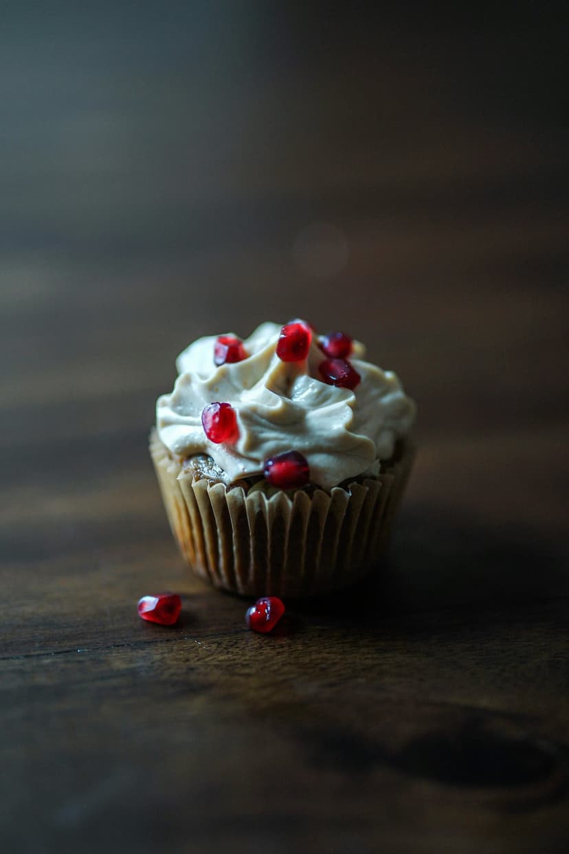 Frosted cupcake with red pomegranate seeds on a brown paper liner atop a dark rustic wooden surface.