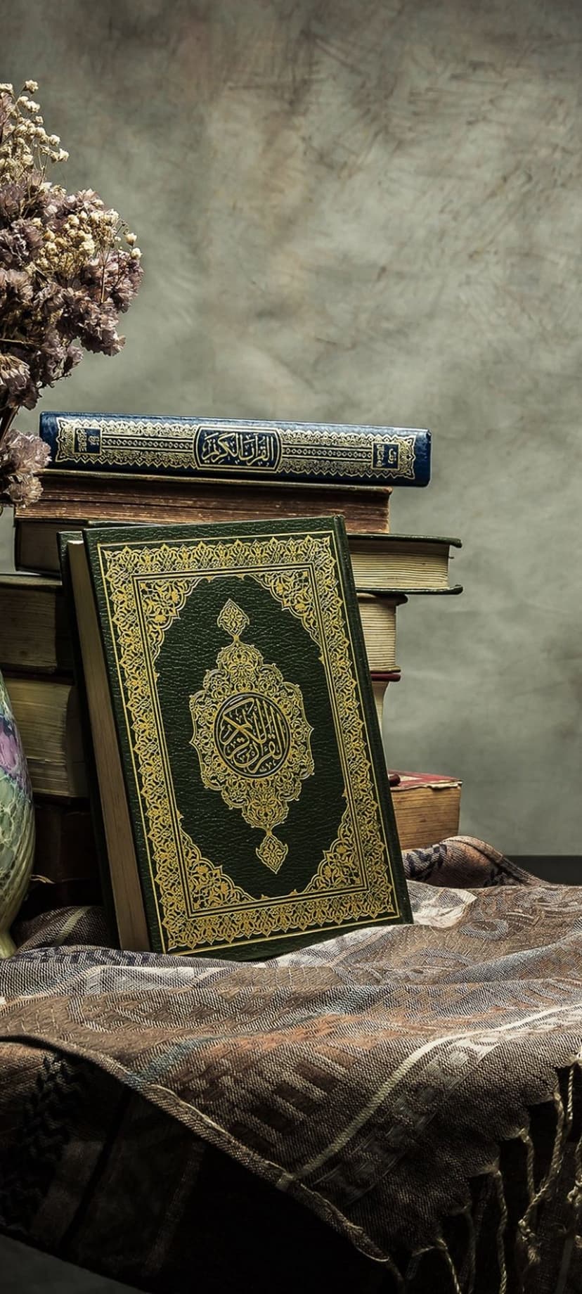 Green Quran with gold accents sits on books beside a vase of dried flowers over a patterned cloth background.