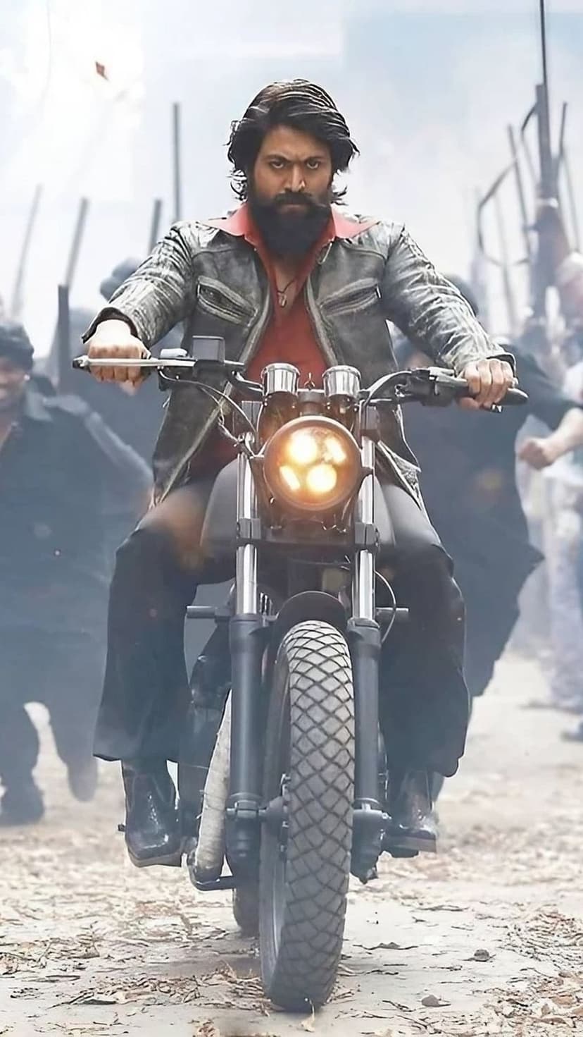 Yash as Rocky Bhai rides a motorcycle wearing a leather jacket and red shirt with a blurred crowd in the background.