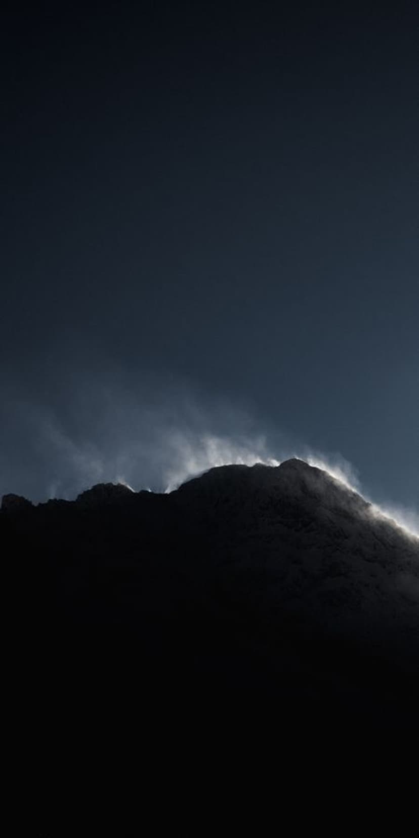 Jagged mountain peak silhouetted against a dark sky with illuminated wisps of fog clinging to the rocky ridge.