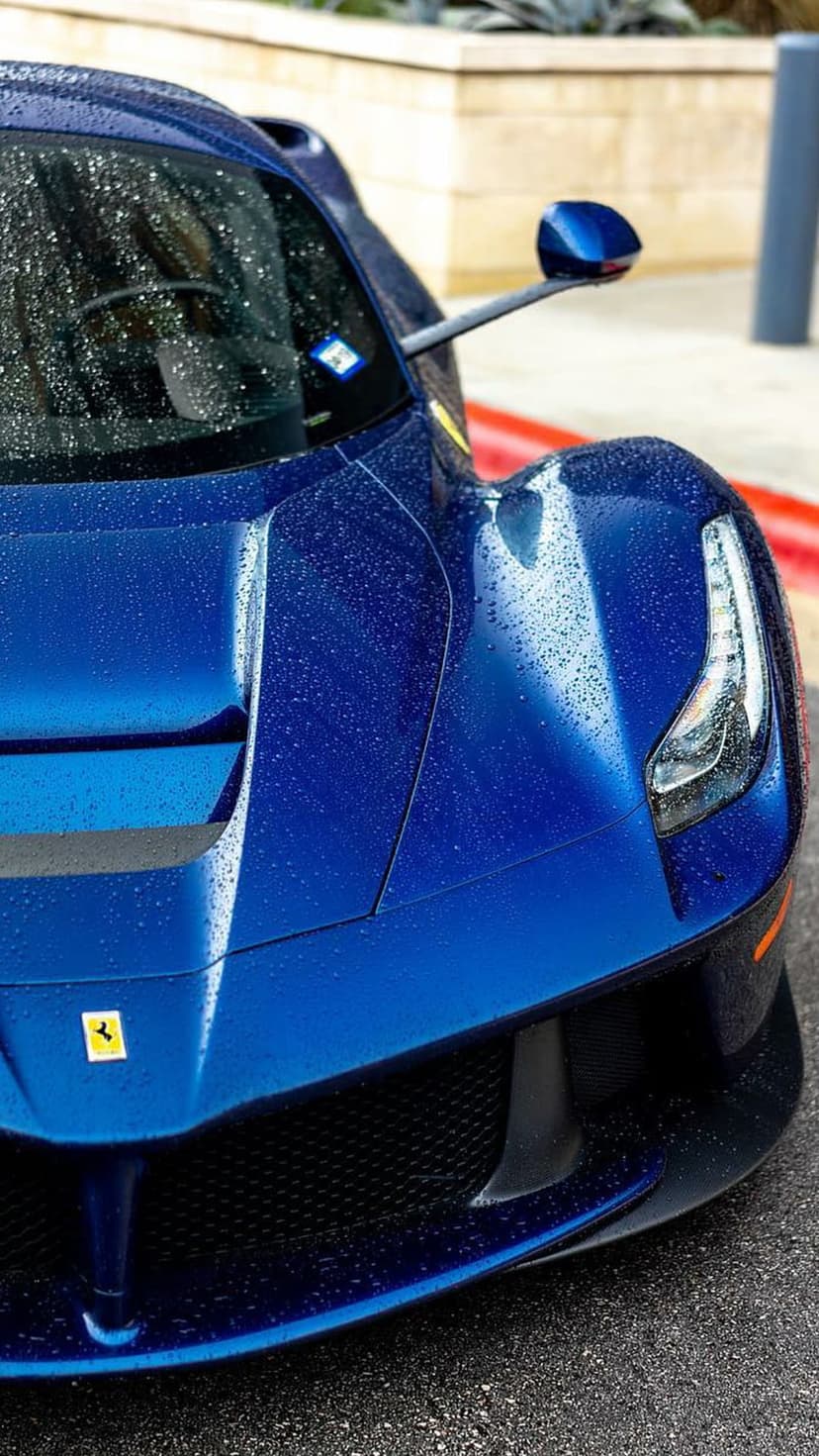 Blue Ferrari supercar covered in glistening raindrops with a close-up on the front emblem and wet hood.