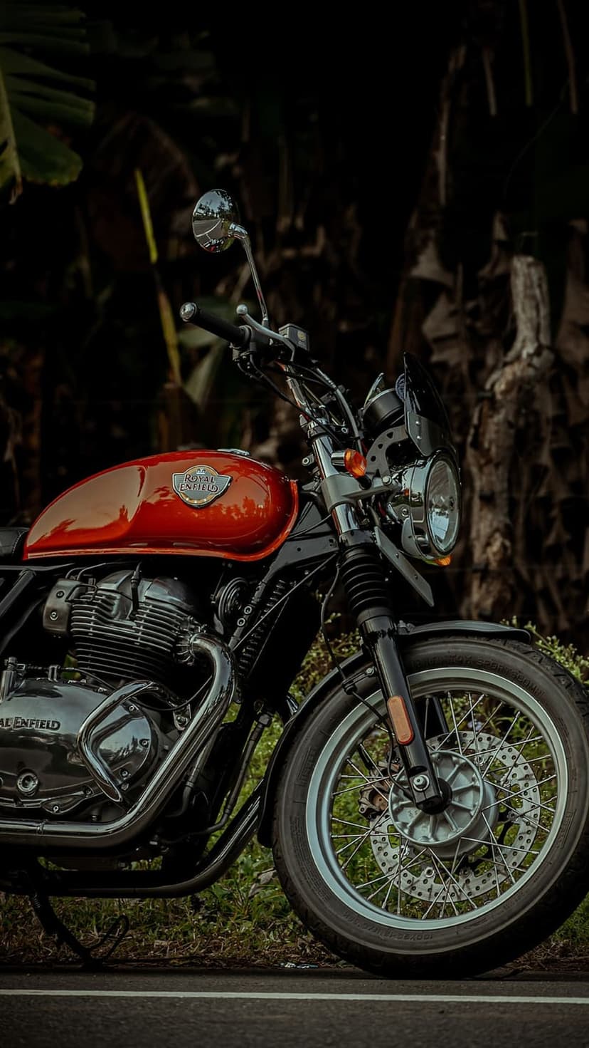 Orange Royal Enfield motorcycle with chrome engine details stands on a roadside against lush green trees.
