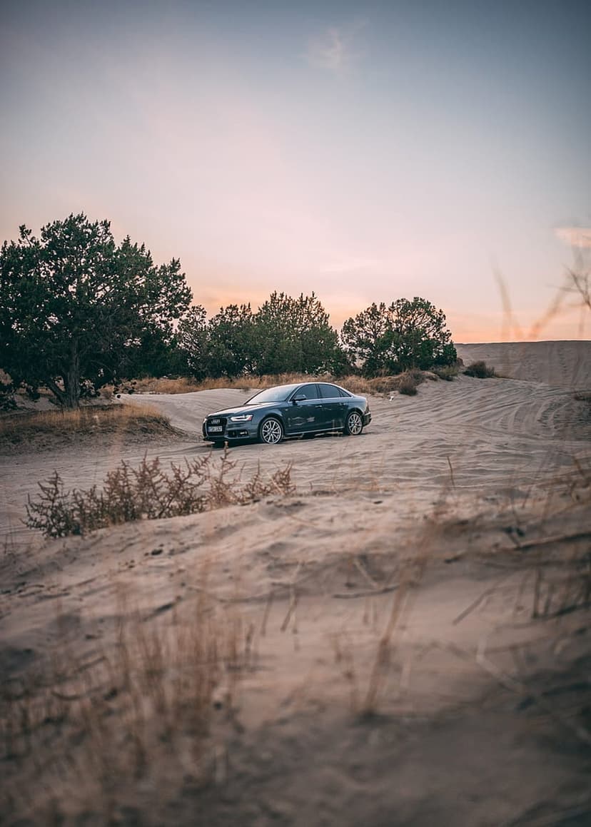 Dark Audi sedan parked on ripples of desert sand dunes under a soft orange and purple sunset sky.