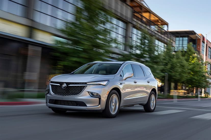Silver Buick Enclave SUV driving past modern glass buildings with motion blur and green trees in daylight.