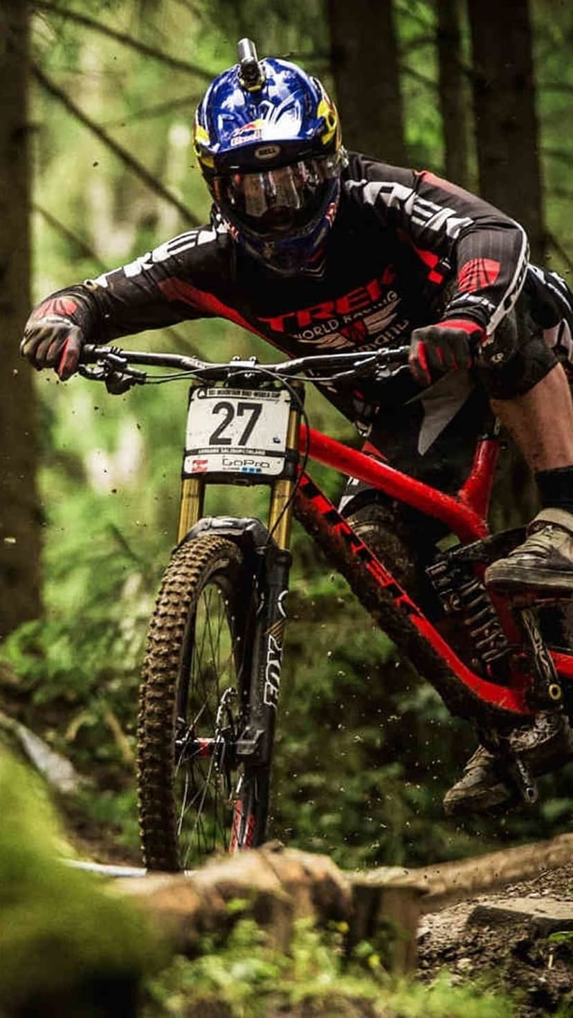 Mountain biker in a full-face helmet leans into a dirt turn while racing through a dense forest trail.