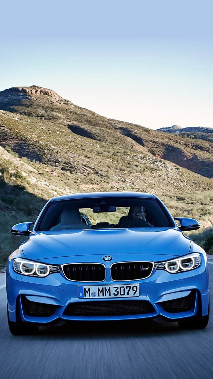 Blue BMW M4 coupe driving on a winding asphalt road through rugged, sun-drenched mountains with headlights glowing.