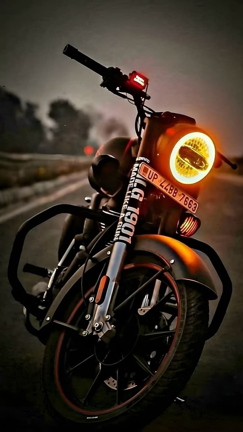 Royal Enfield motorcycle with a glowing yellow headlight and red indicator parked on a dark road at dusk.