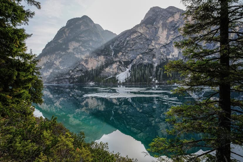 Jagged mountains covered in mist reflect in a calm emerald lake framed by dark green evergreen trees.