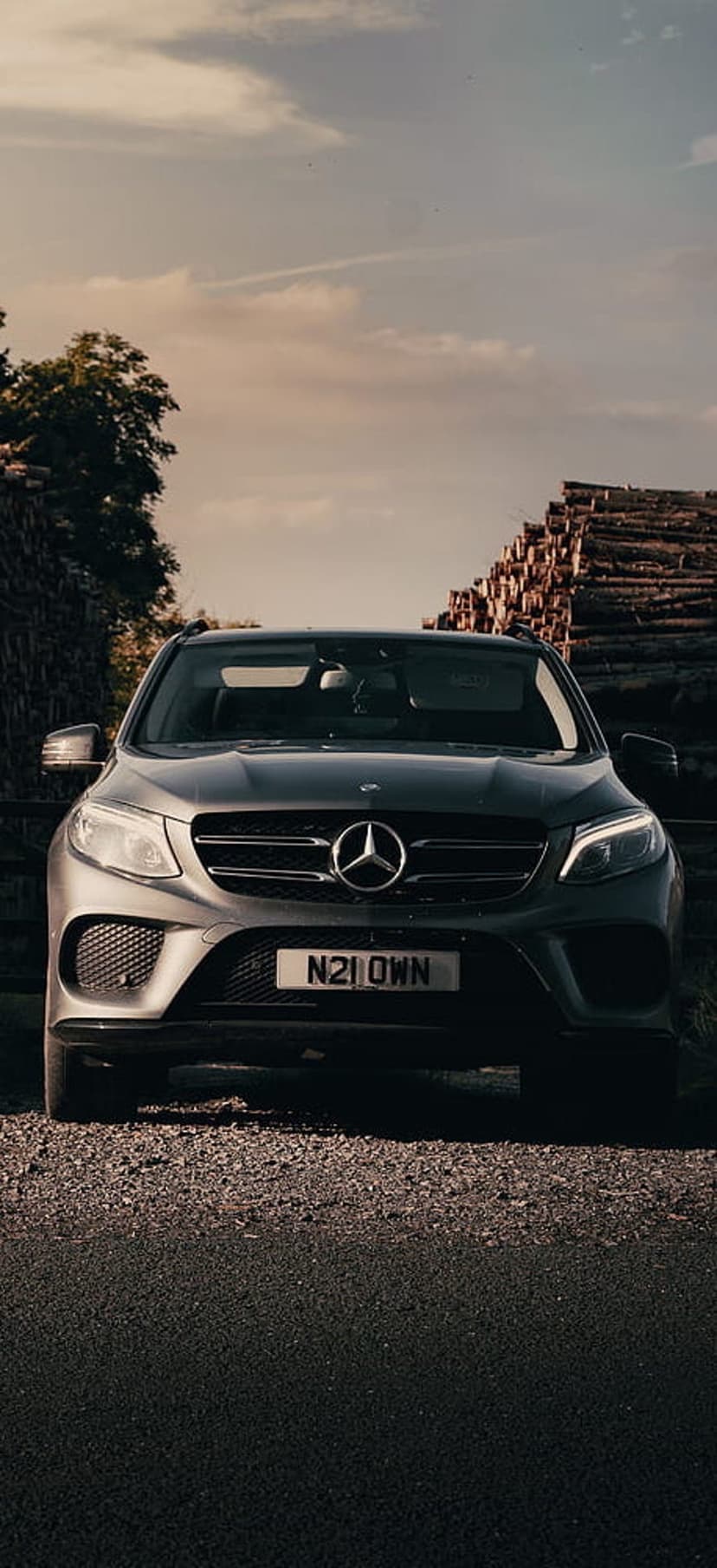 Mercedes-Benz GLE Coupe parked on gravel beside a wall of logs under a moody sunset with glowing headlights.
