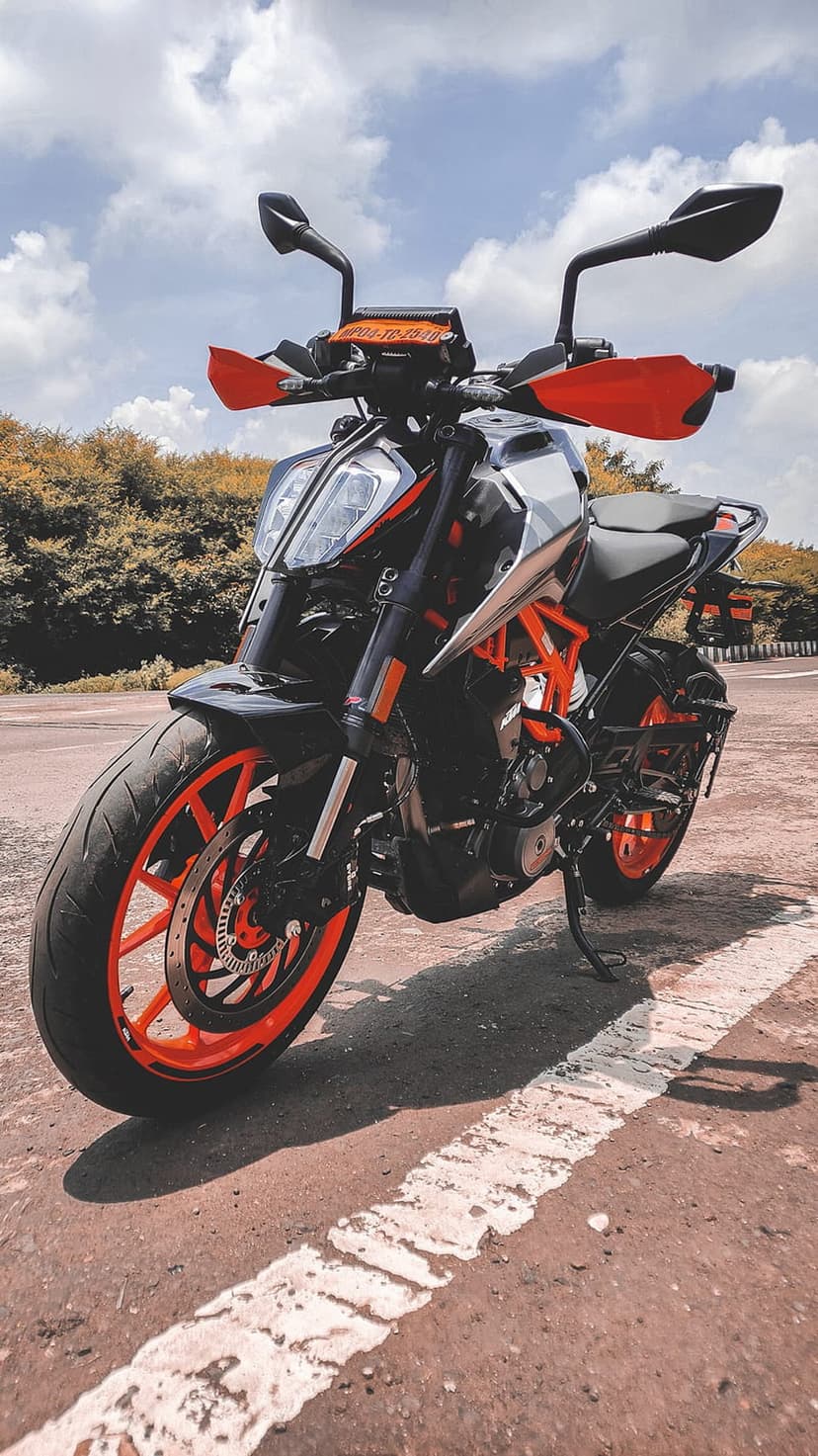KTM Duke motorcycle with orange rims and black bodywork parked on a paved road near a lush green forest line.