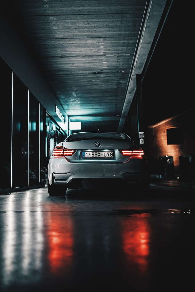 Silver BMW M4 sports car with glowing red taillights parked on a wet floor inside a modern building at night.