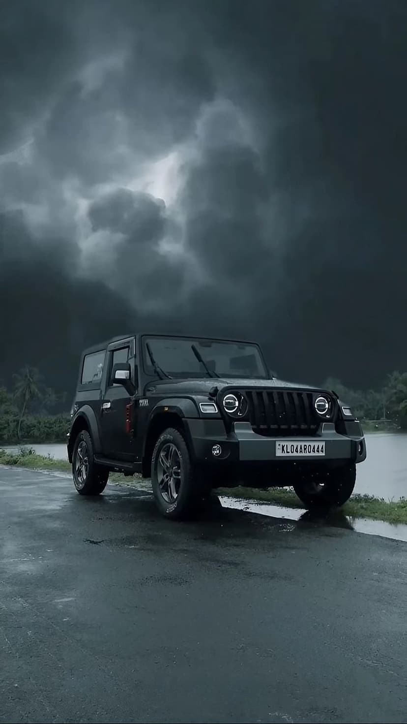 Black Mahindra Thar SUV parked on a wet road with reflections under a dark stormy sky and green trees.