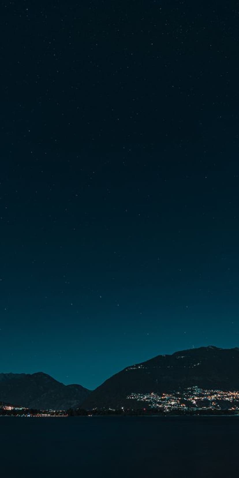 Glowing village lights reflect in a dark lake beneath a vast starry sky and distant mountain silhouettes.