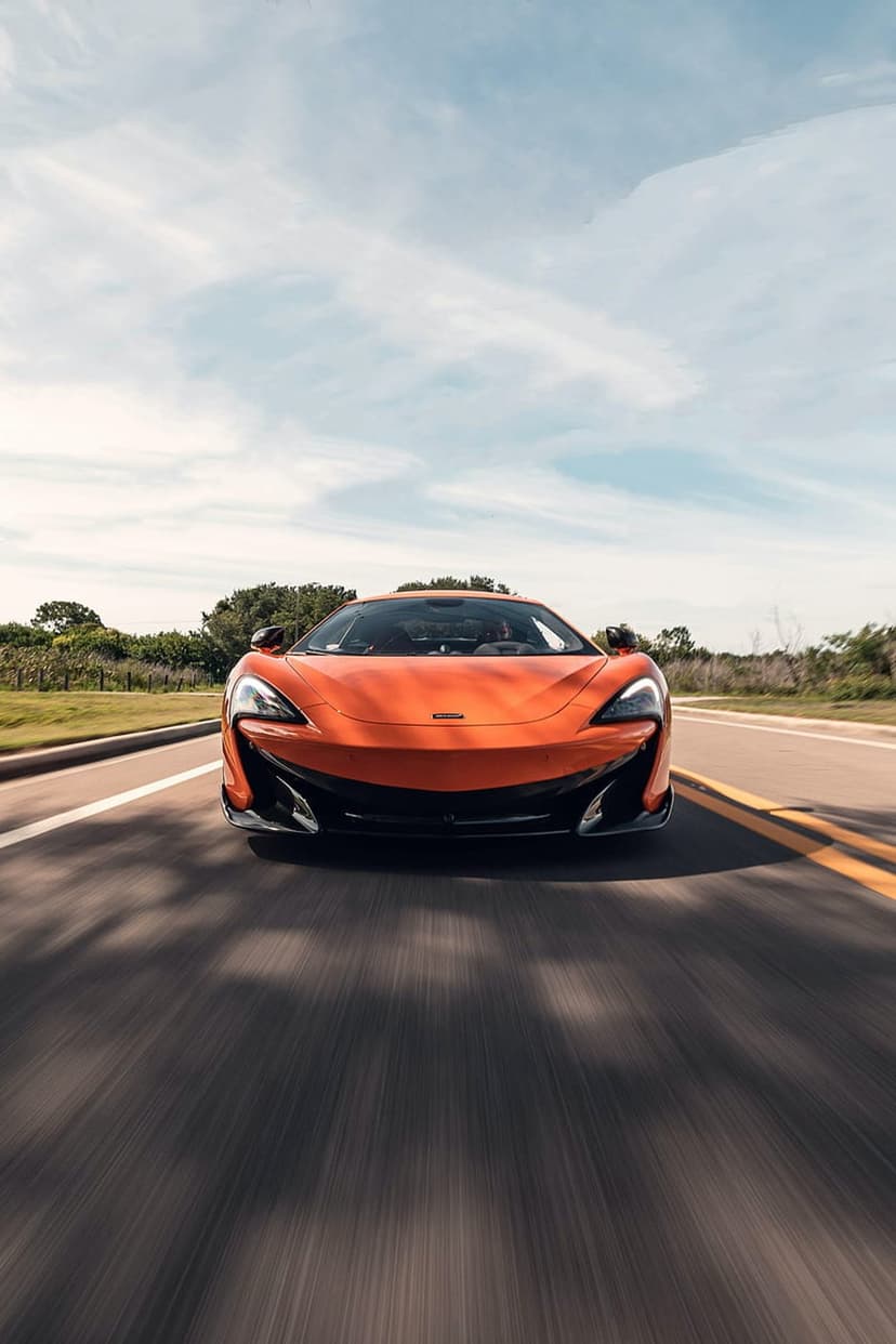 Orange McLaren supercar speeds along a tree-lined road under a bright sky with visible motion blur effects.