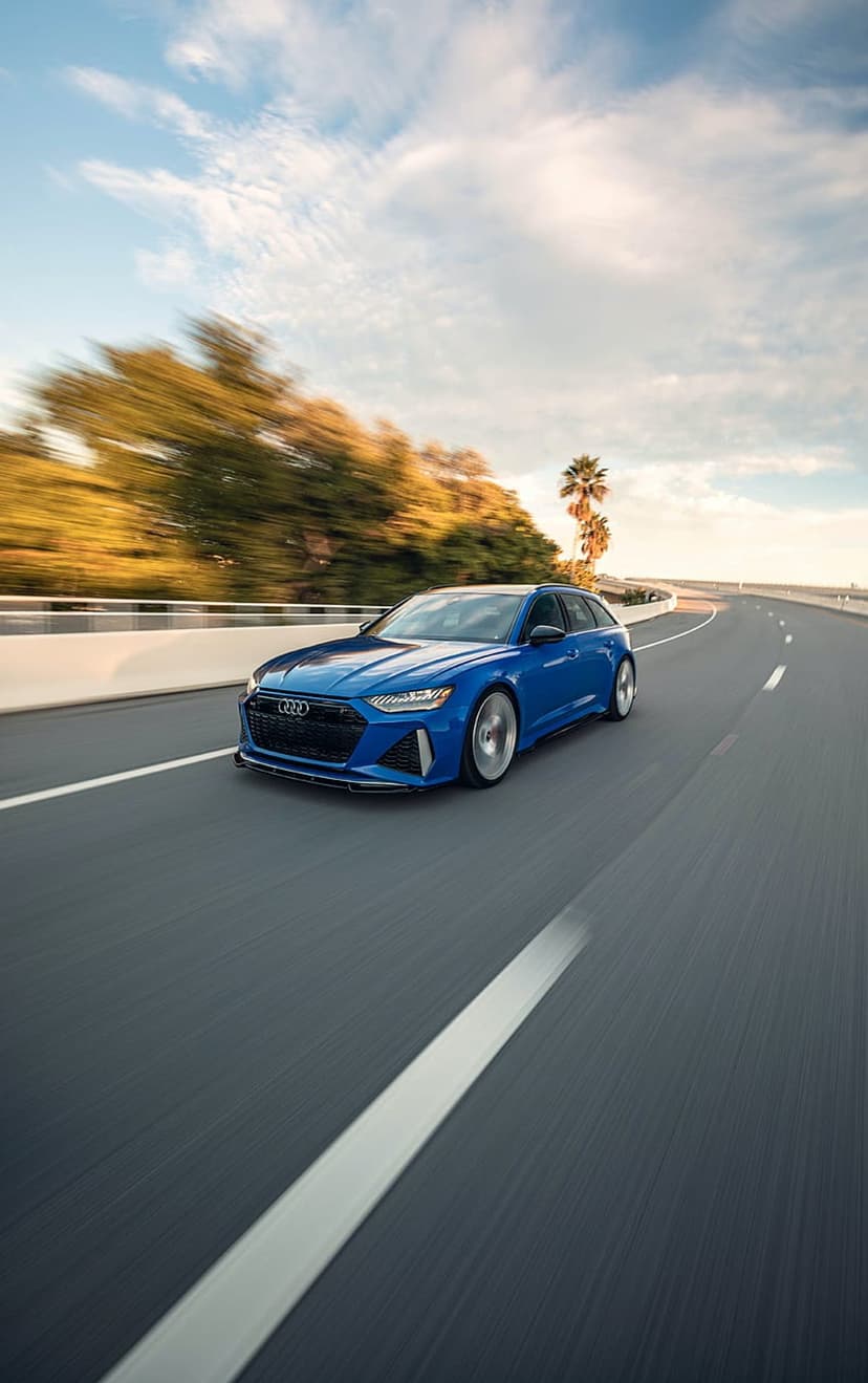 Blue Audi RS6 Avant sports wagon driving on a paved road lined with green trees and palms under a bright sky.