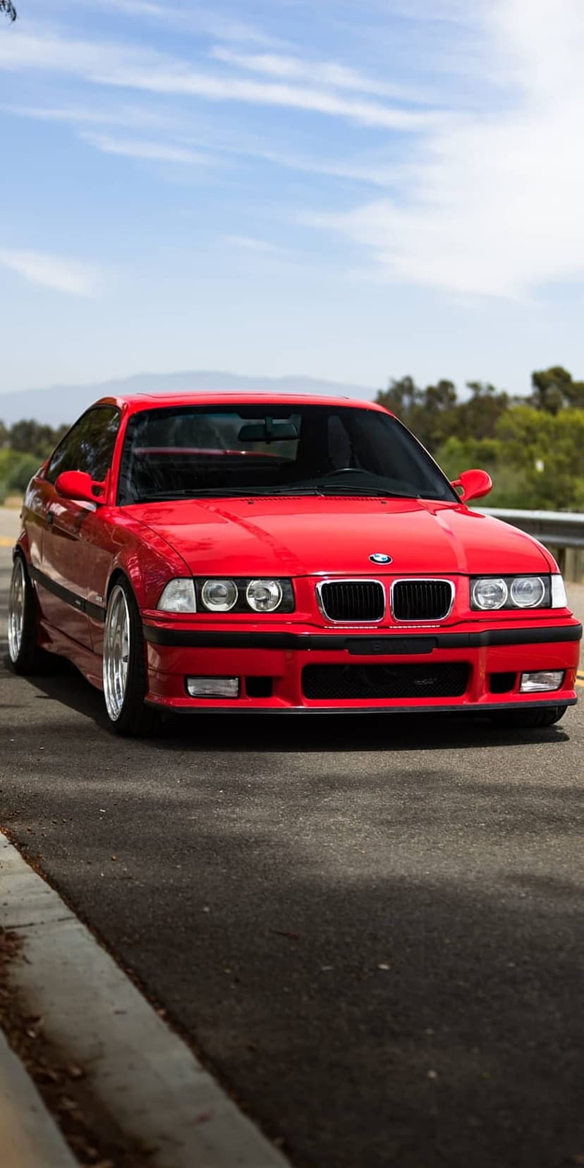 Red BMW E36 coupe drives along a paved road under a blue sky with green trees and hills in the background.