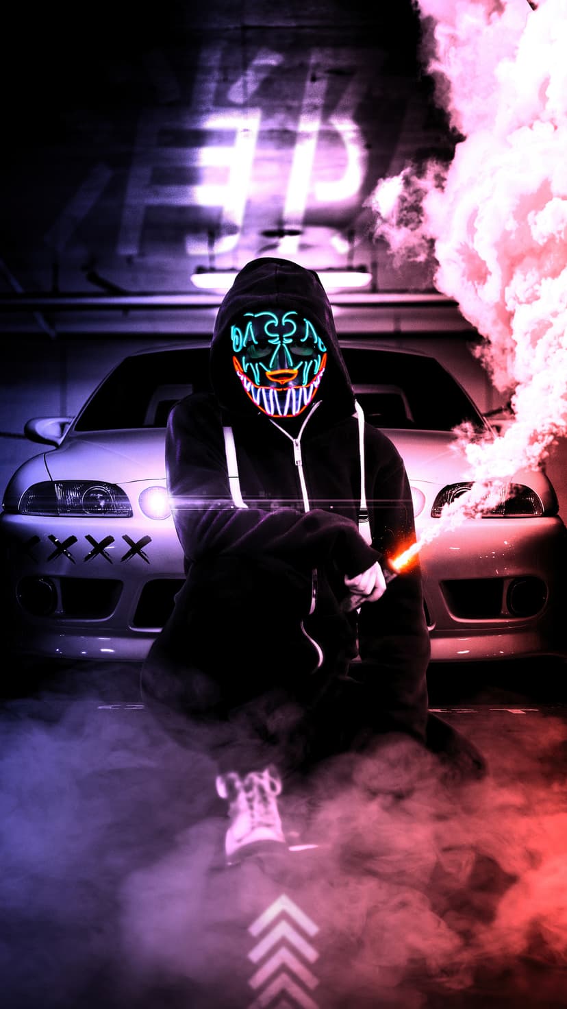 Masked person in a fanged LED mask crouches near a white car while surrounded by pink and white smoke clouds.