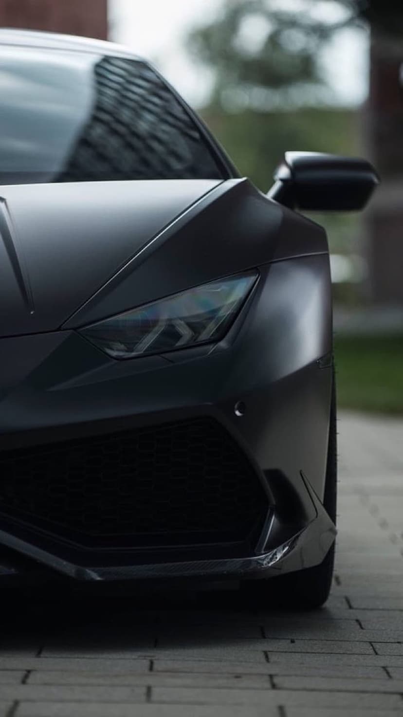 Matte black Lamborghini Huracan front grille and LED headlights on a paved road with blurred greenery.