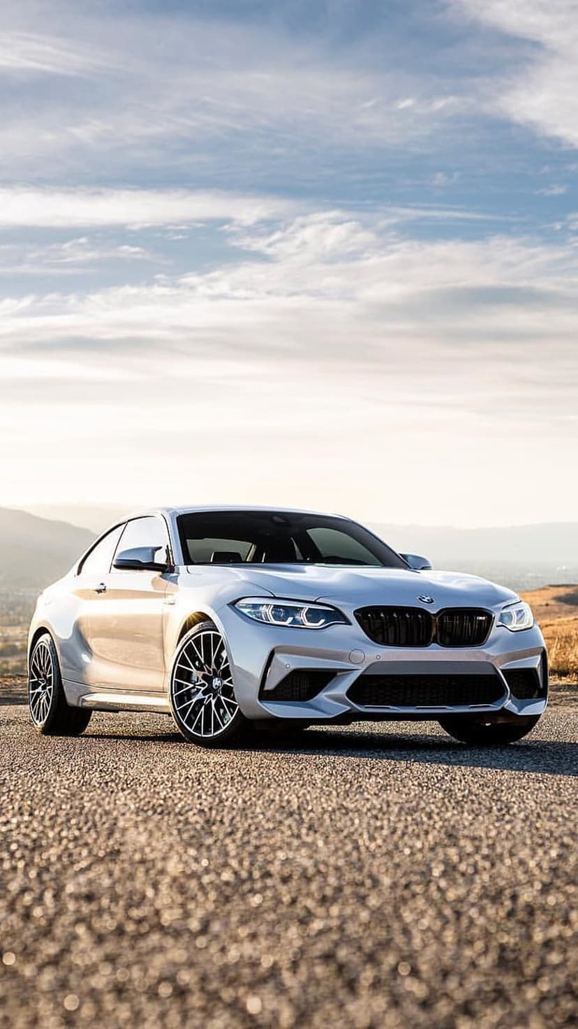 Silver BMW M2 Coupe stationary on dark pavement with misty ridges and grey clouds in the distance.