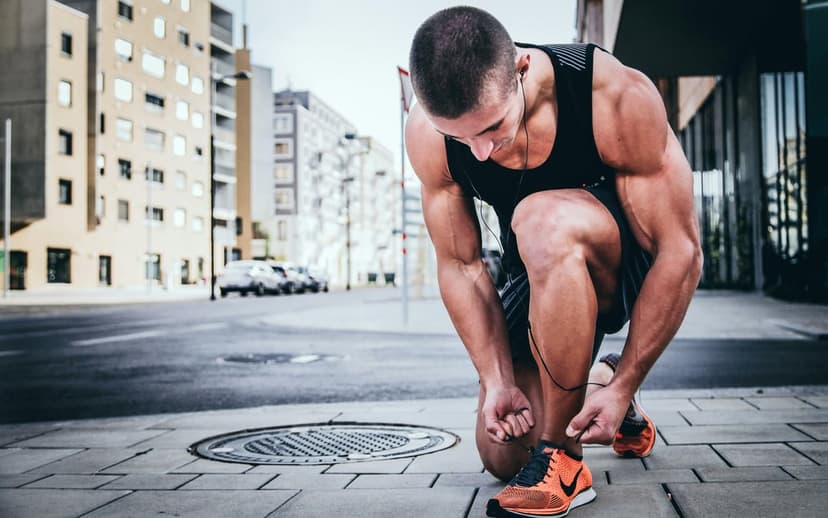 Muscular man in a black tank top ties orange running shoes on a city sidewalk with buildings in the background.