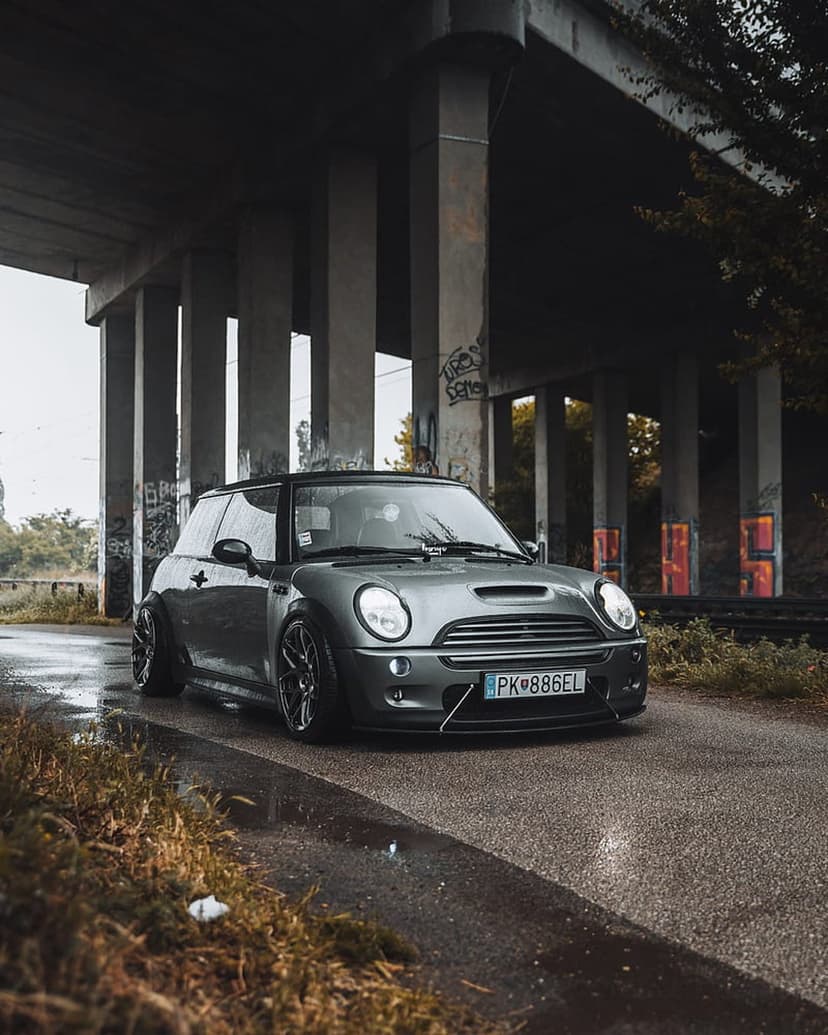 Grey Mini Cooper S parked on wet asphalt under a concrete bridge with graffiti and industrial pillars.