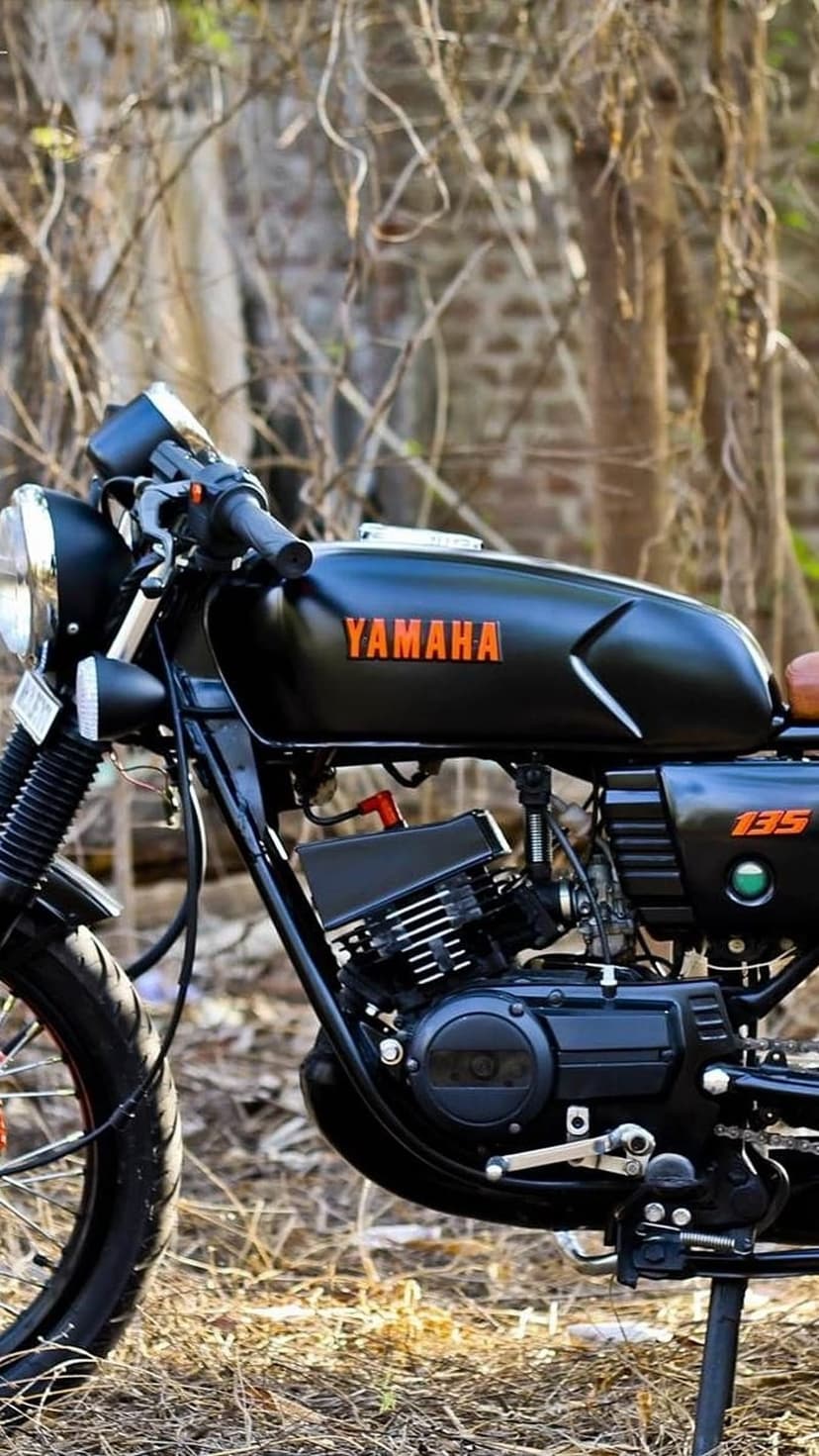 Yamaha RX 135 motorcycle with matte black paint and orange logo parked among dry autumn leaves and forest trees.