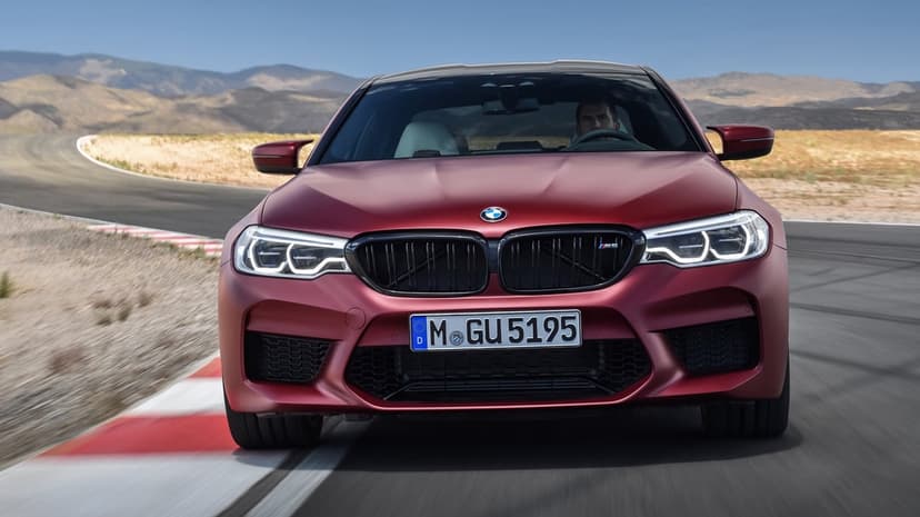 Matte red BMW M5 racing on a curved asphalt track with red and white curbs under a clear blue desert sky.