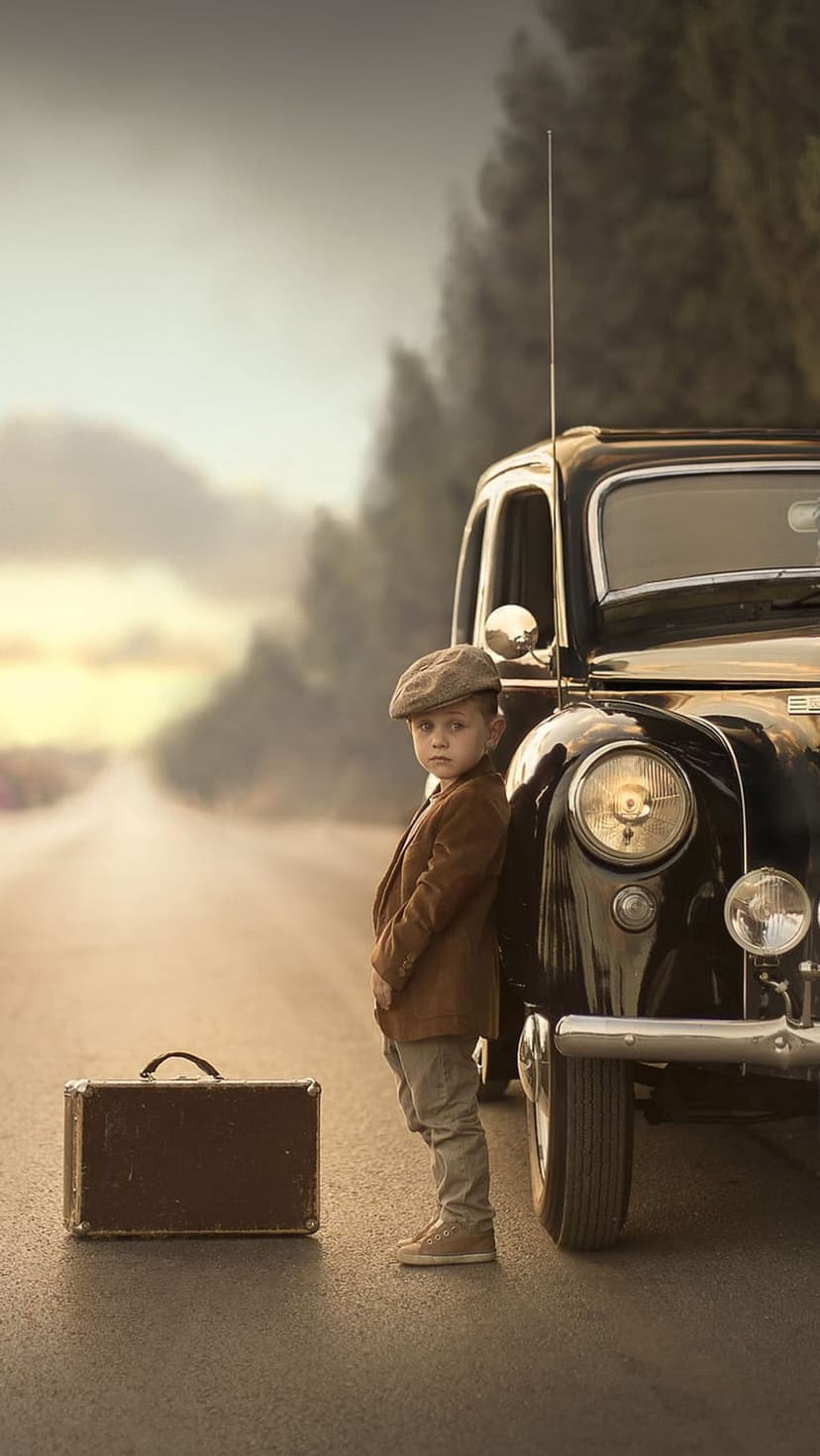 Young boy in a flat cap stands by a black vintage car and suitcase on a hazy, golden country road.