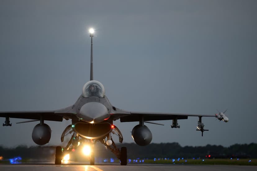 F-16 fighter jet with bright landing lights facing forward on an asphalt runway under a dark twilight sky.