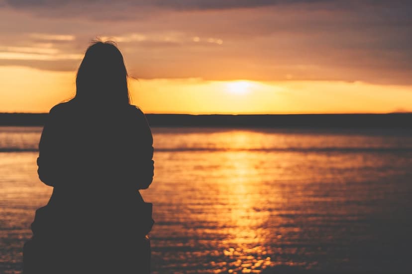 Woman's dark silhouette overlooks a calm sea beneath a vibrant orange and purple twilight sky during sunset.