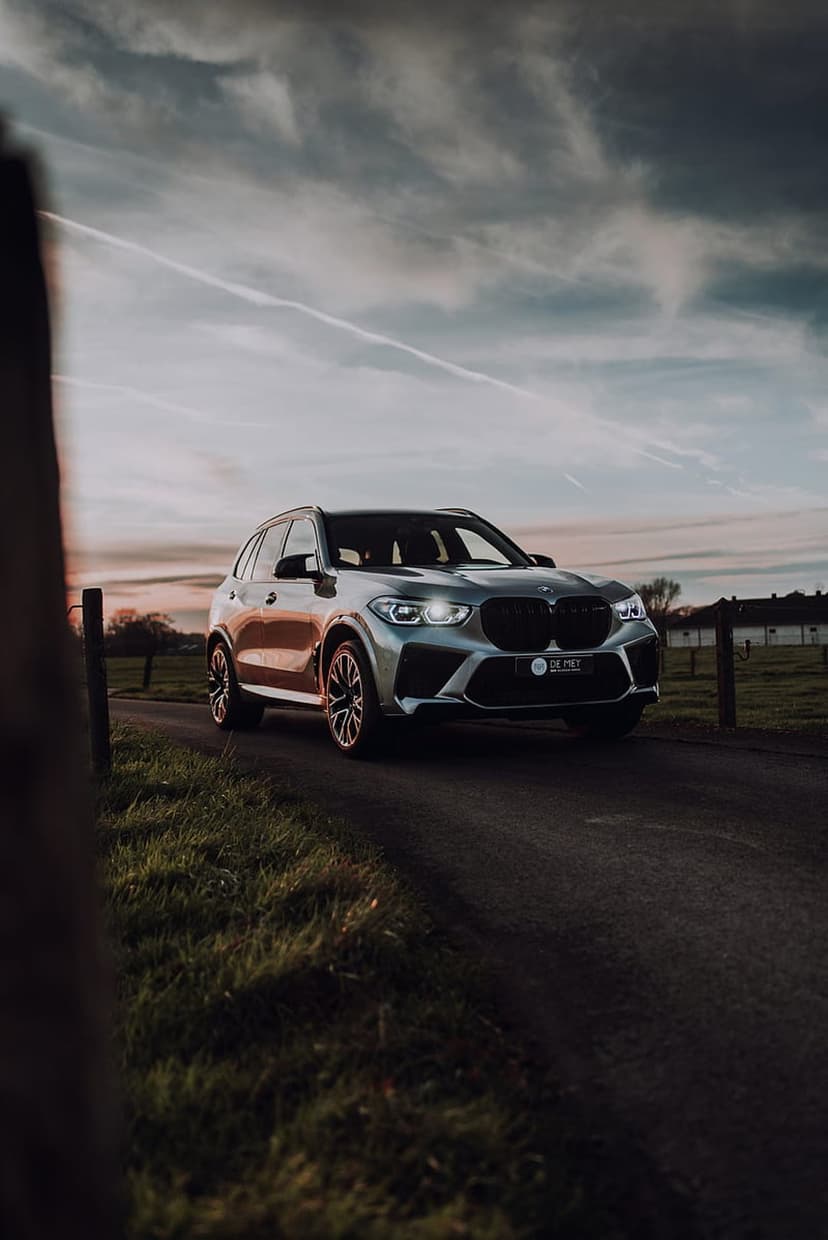 Silver BMW SUV with glowing headlights drives on a rural road during a vibrant sunset with wispy cloud formations.