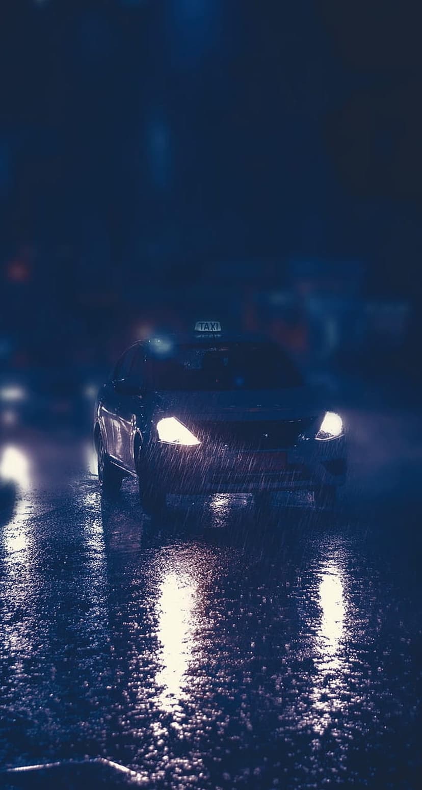 Yellow taxi cab driving through heavy rain at night with bright headlights and glowing roof sign reflections.