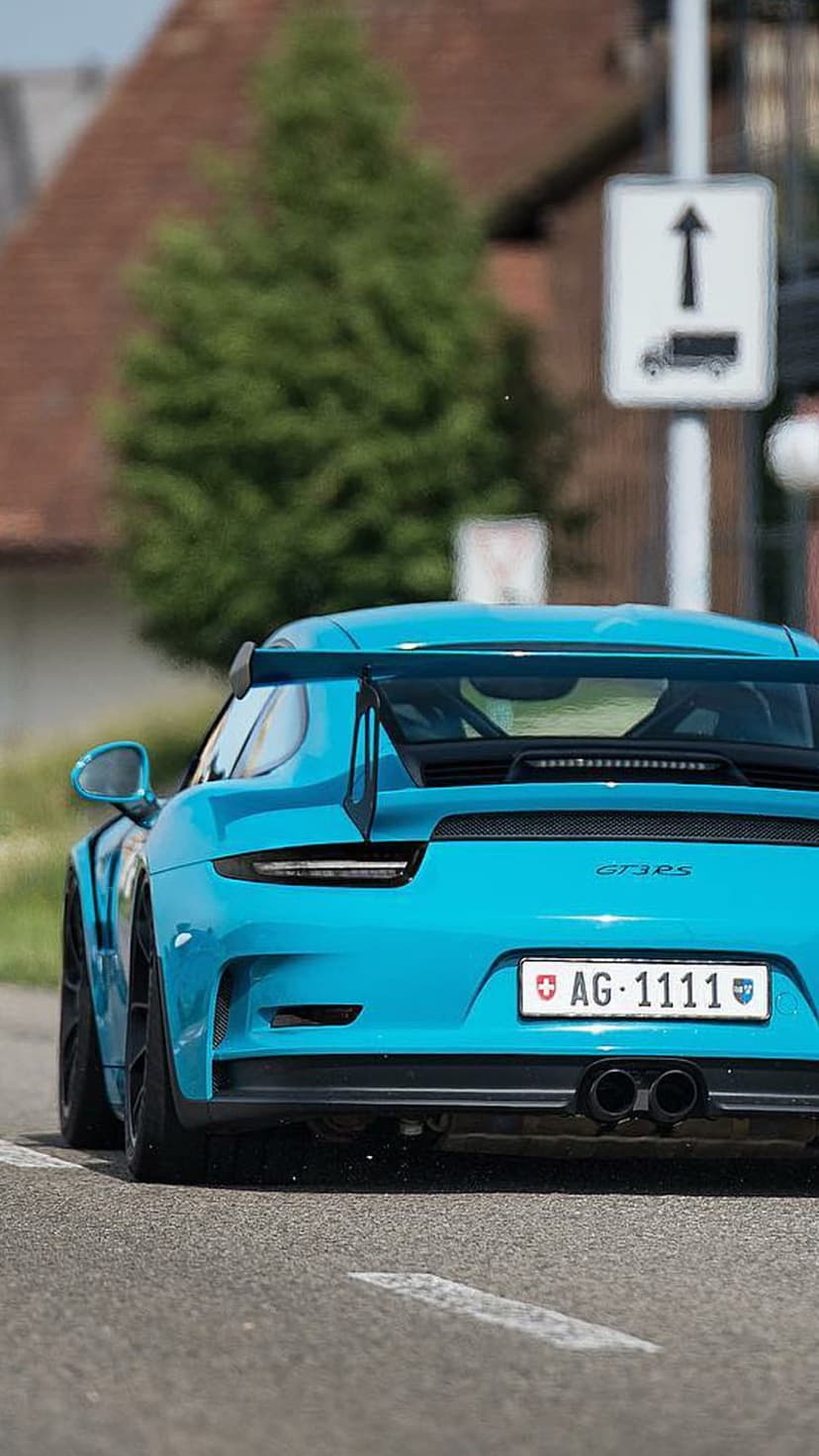 Blue Porsche 911 GT3 RS with a large rear wing speeds past a green tree and traffic sign on a blurred road.