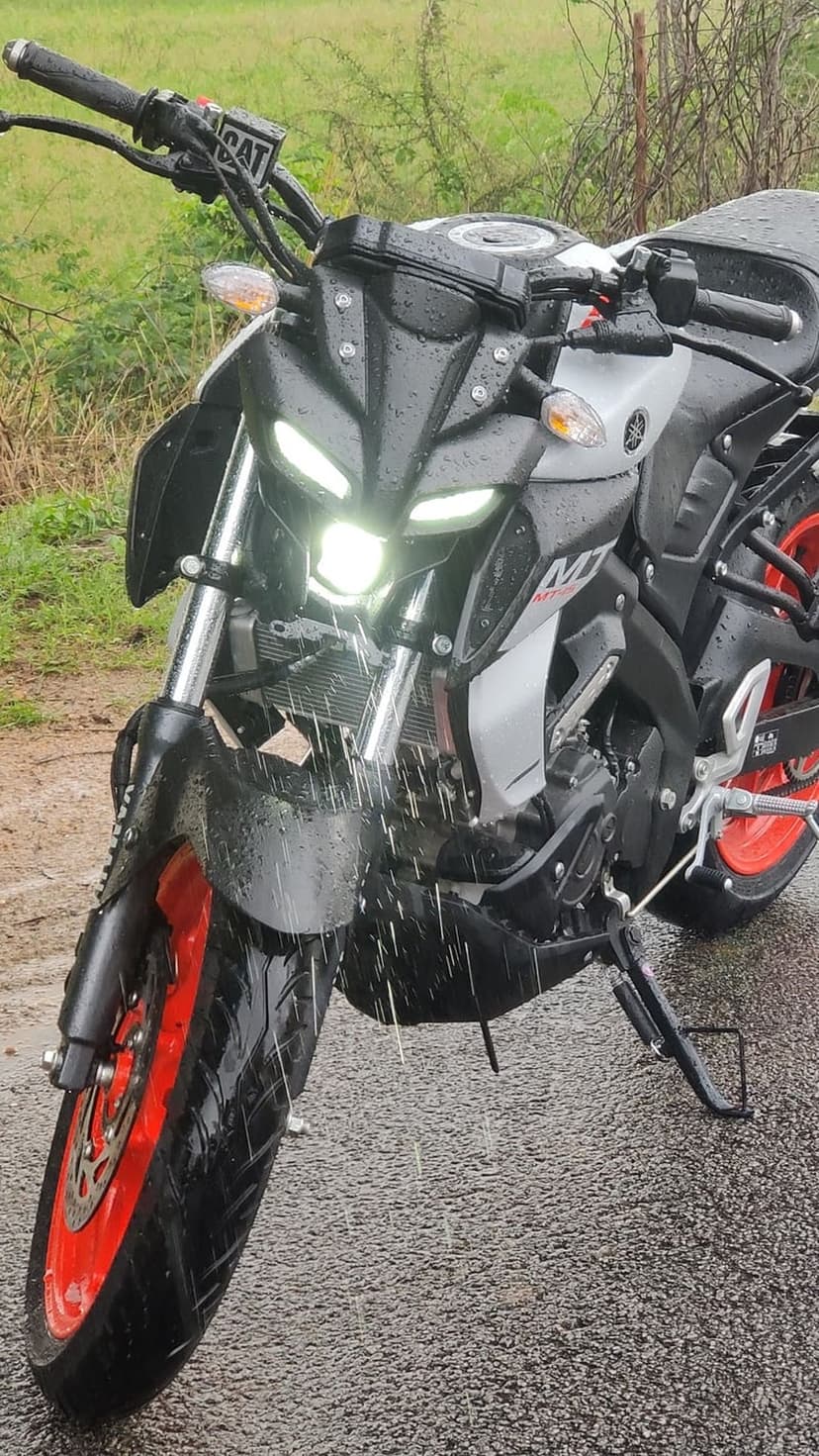 Yamaha MT-15 motorcycle with orange wheels parked on wet asphalt under falling rain during a moody evening.