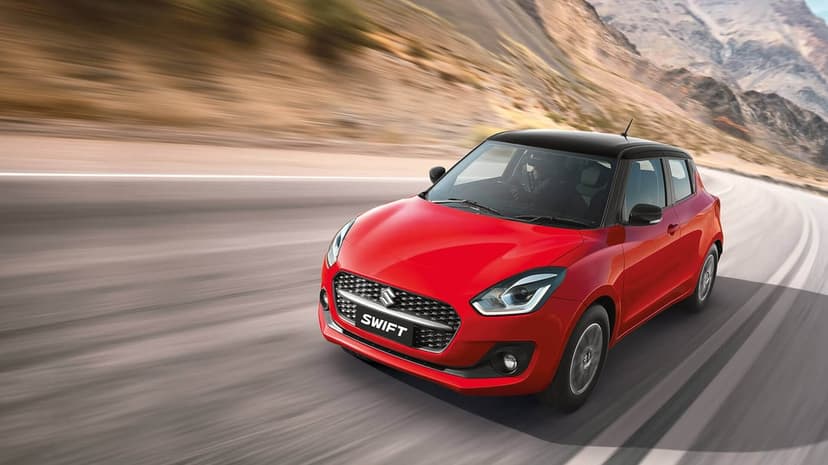 Red Suzuki Swift hatchback with a black roof driving on a mountain road with motion blur background.