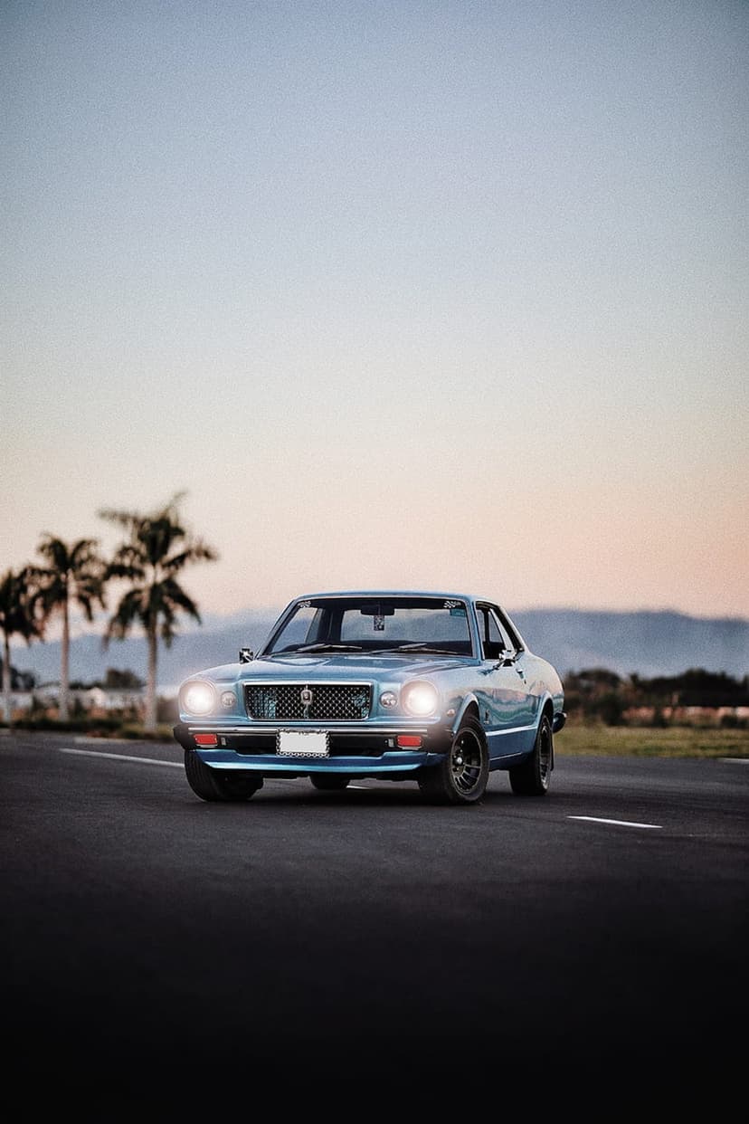 Vintage blue coupe driving on a coastal road lined with palm trees under a vibrant orange and purple sunset sky.