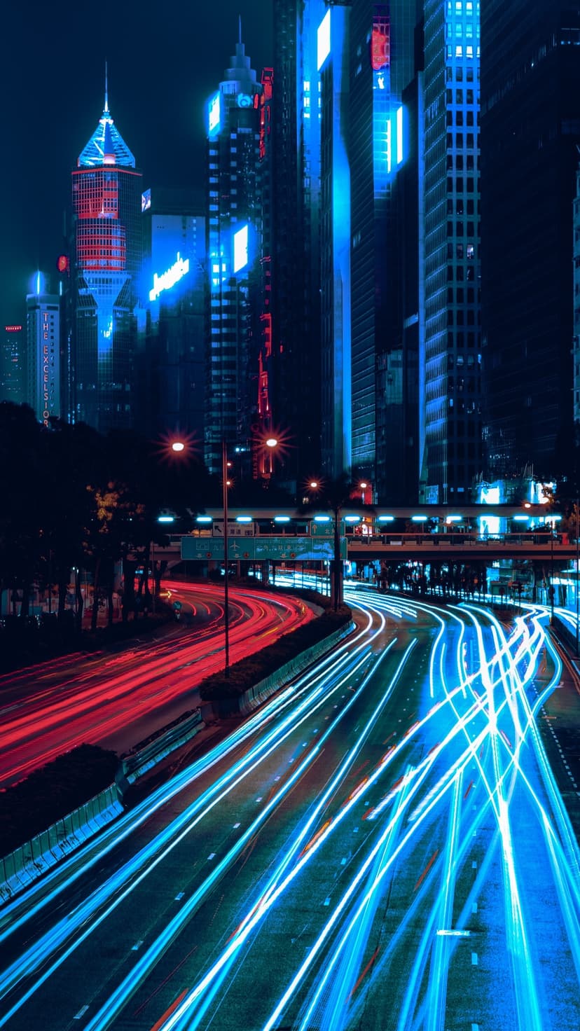 Red and blue light trails flow along a vertical highway between glowing neon skyscrapers in a night city scene.